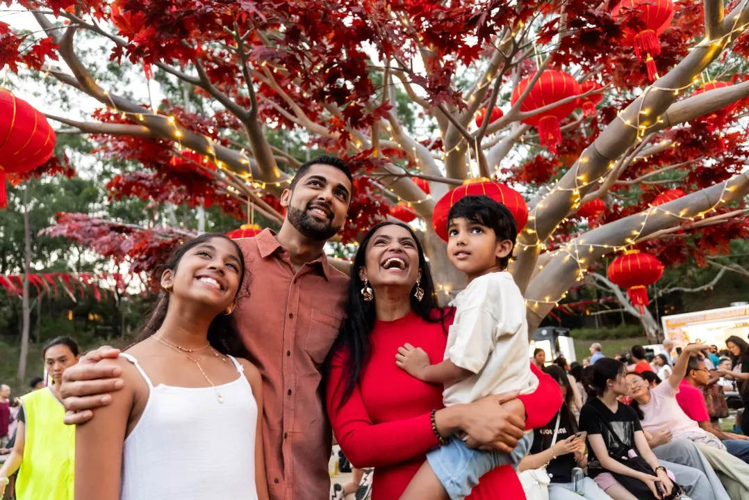 A joyful family of four stands under a tree adorned with red lanterns. The parents smile, the mother holds a young child, and a teenage girl grins beside them.