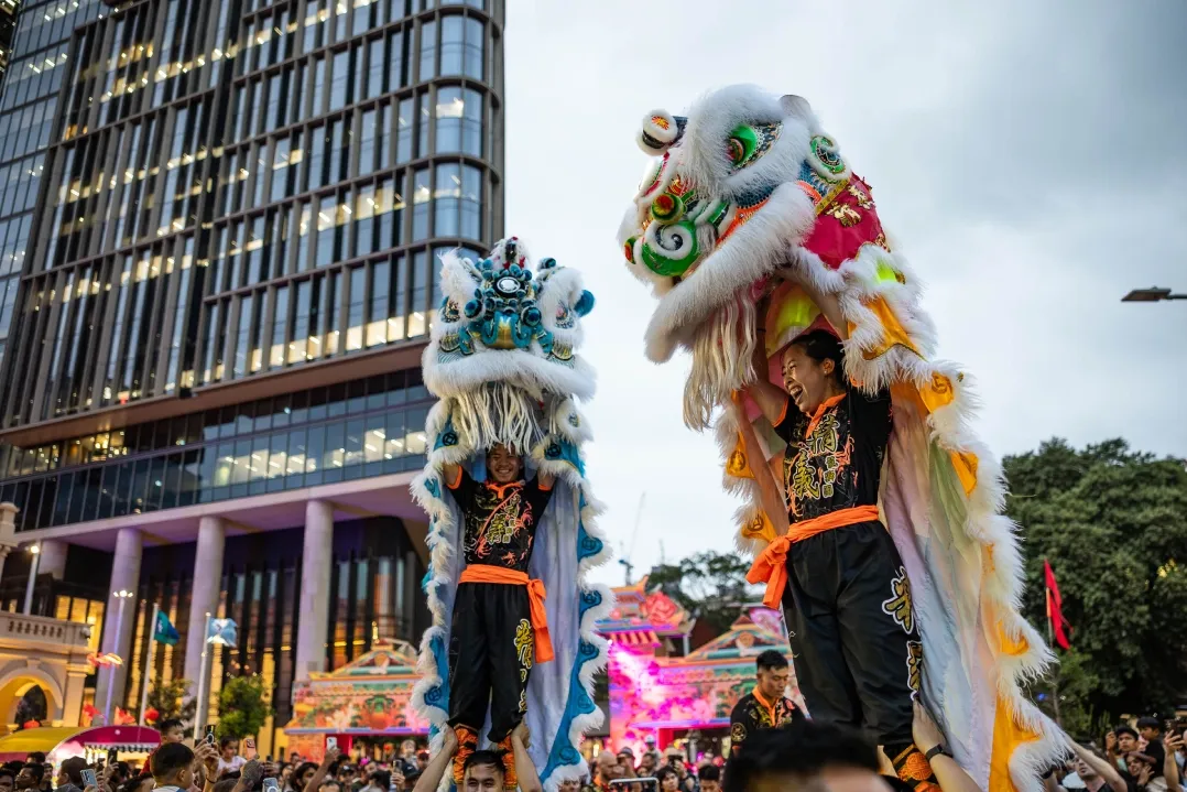 Lion dancers in vibrant costumes perform energetically on a street against the backdrop of a modern building, evoking a festive and lively atmosphere.