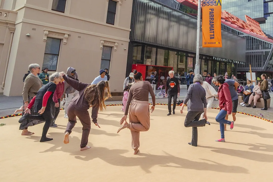 A group of people in colorful traditional attire energetically perform a dance on a sandy street, with onlookers watching near modern buildings.