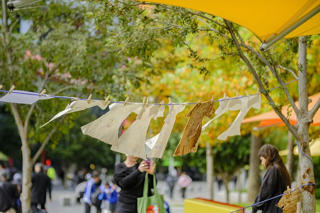 People strolling under vibrant yellow umbrellas alongside a line of trees in a lively outdoor market setting, conveying a cheerful and bustling atmosphere.