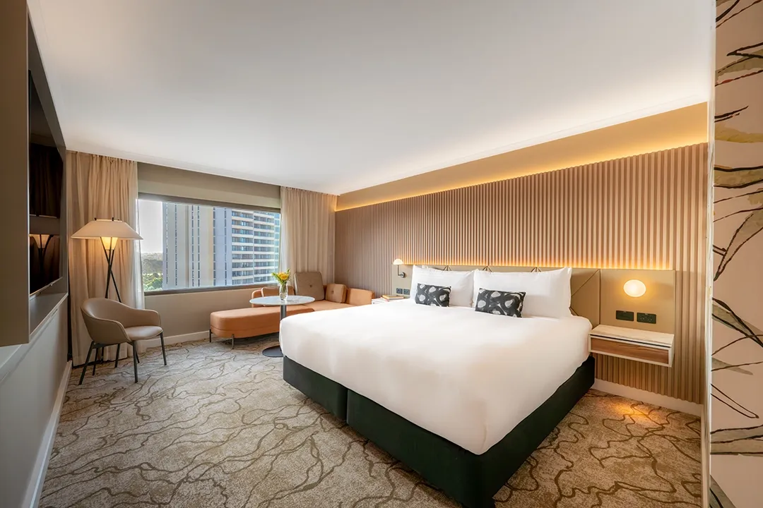 Modern hotel room with a king-size bed, white linens, cushioned headboard, warm lighting, cozy seating area by a window, and a scenic city view.