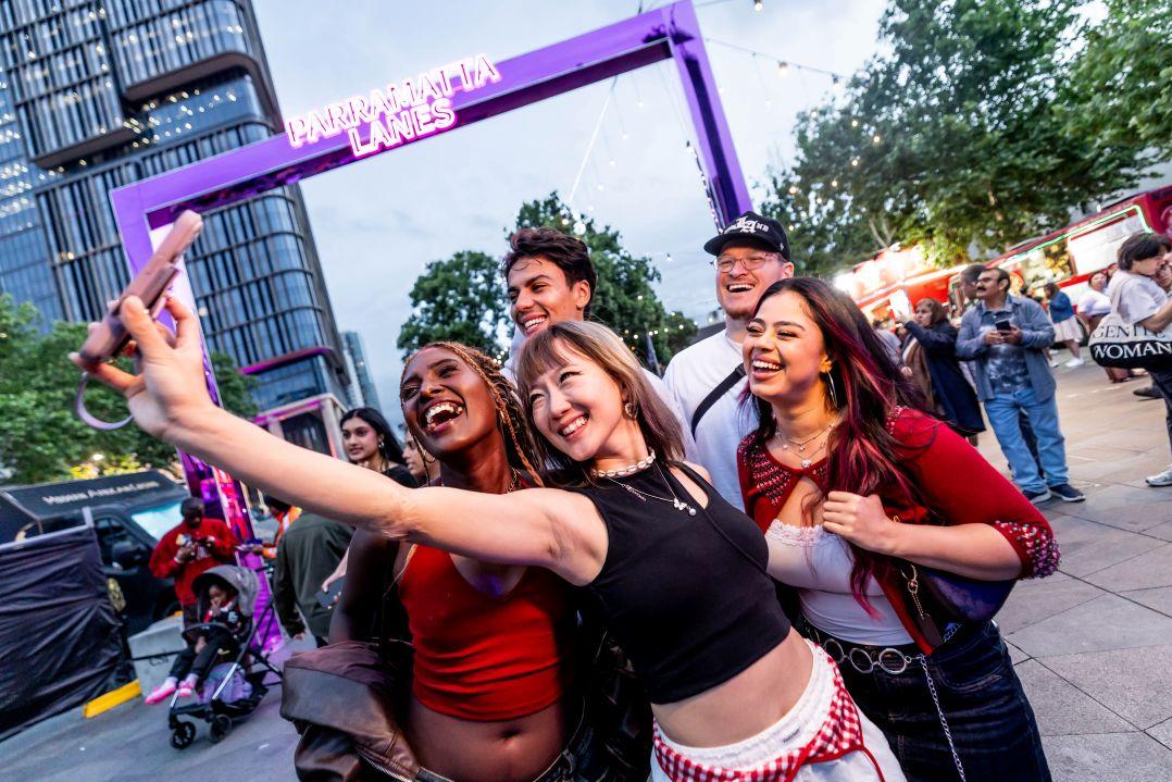 A group of five people happily take a selfie outdoors under a purple frame. They are smiling and the scene exudes a lively, fun atmosphere.