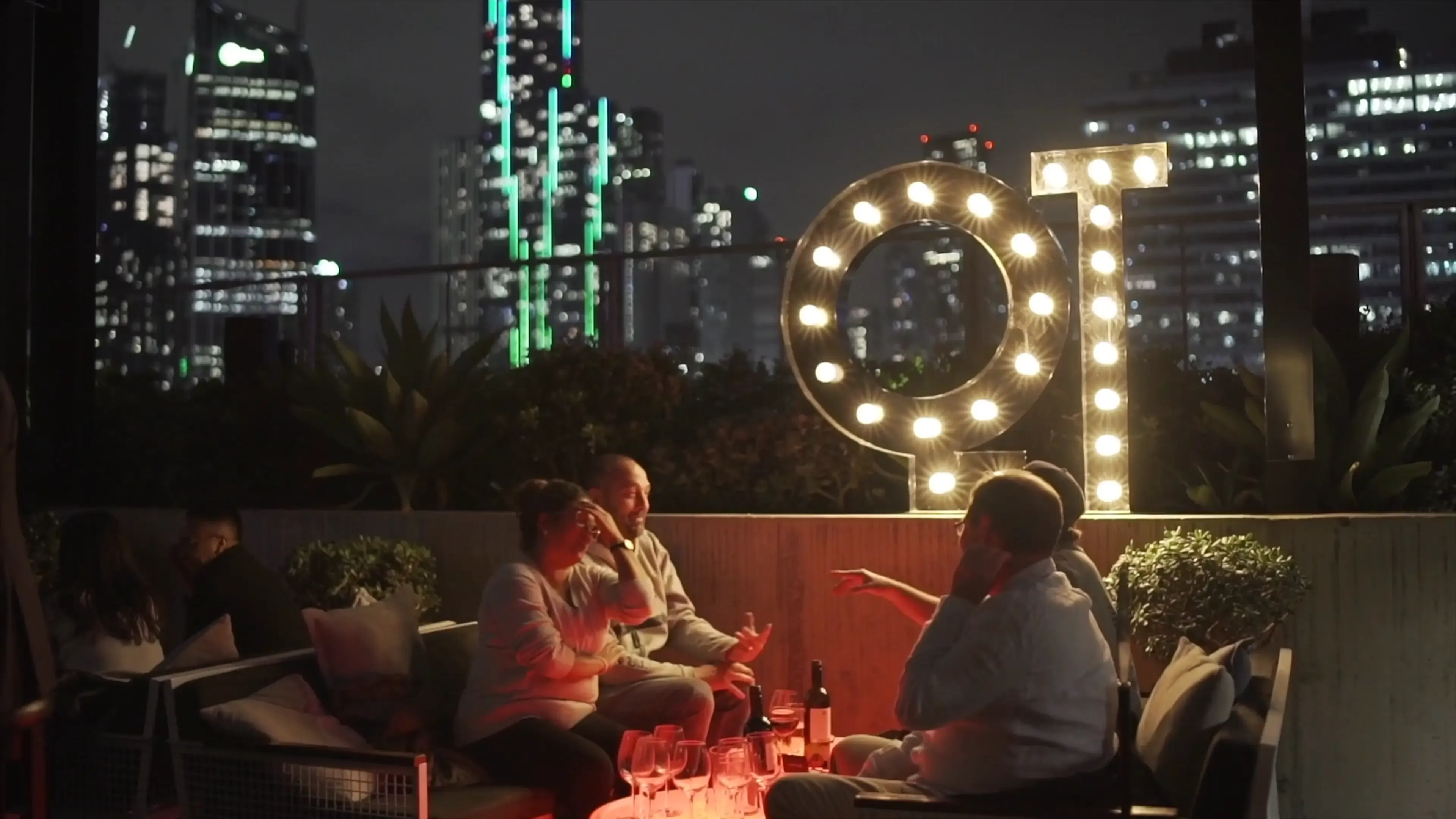 A rooftop bar scene at night shows three people conversing around a table with drinks. A lit "QT" sign glows warmly, and city skyscrapers loom in the background.