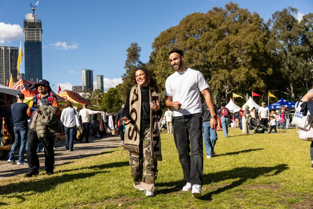 A joyful couple poses on a sunny day at a park event with a lively crowd, food stalls, and city skyline in the background, conveying a festive atmosphere.