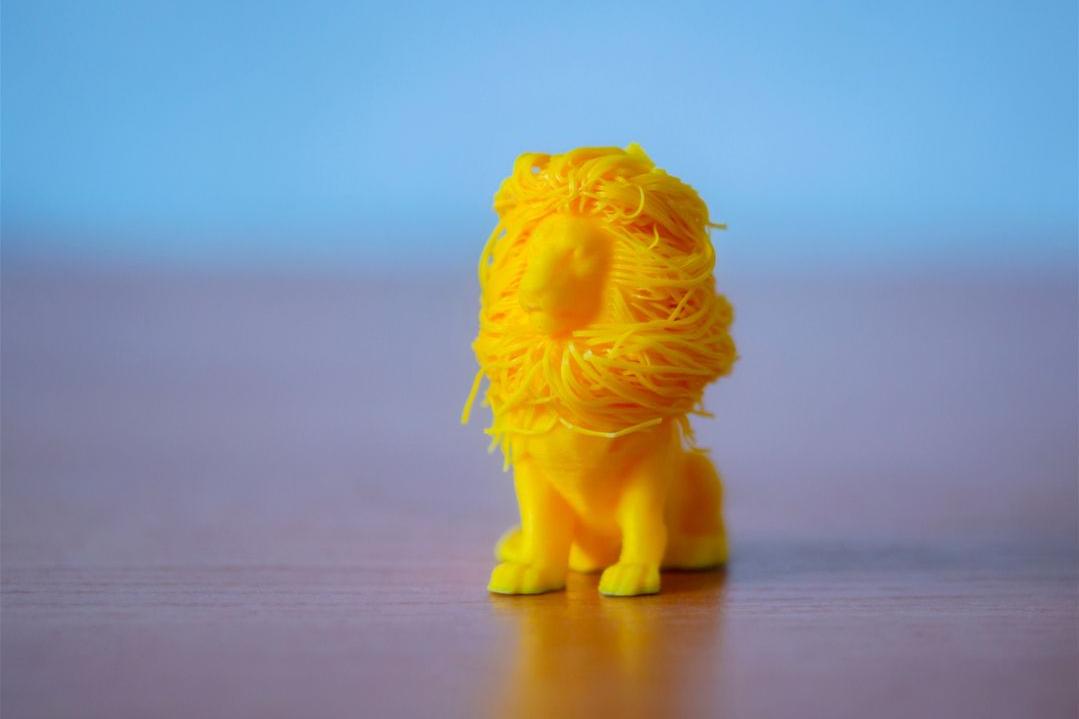 A small yellow 3D-printed lion with a tangled, intricately textured mane sits on a wooden surface against a soft blue background.
