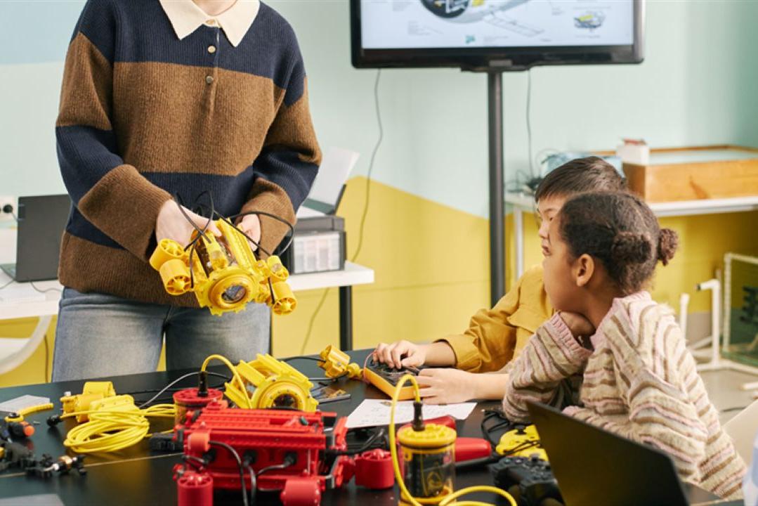 A teacher shows robotic parts to three attentive students at a table with electronics. The classroom setting conveys a sense of curiosity and learning.