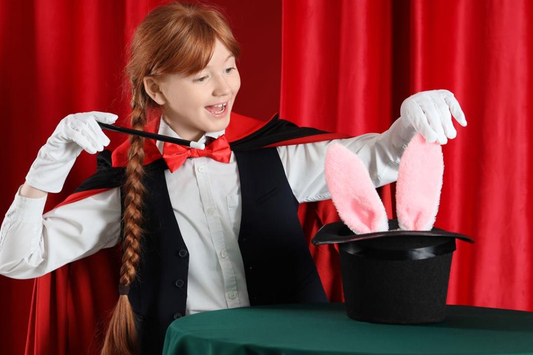 A smiling girl in magician attire with a red cape and gloves uses a wand over a top hat with pink bunny ears, set against red curtains.