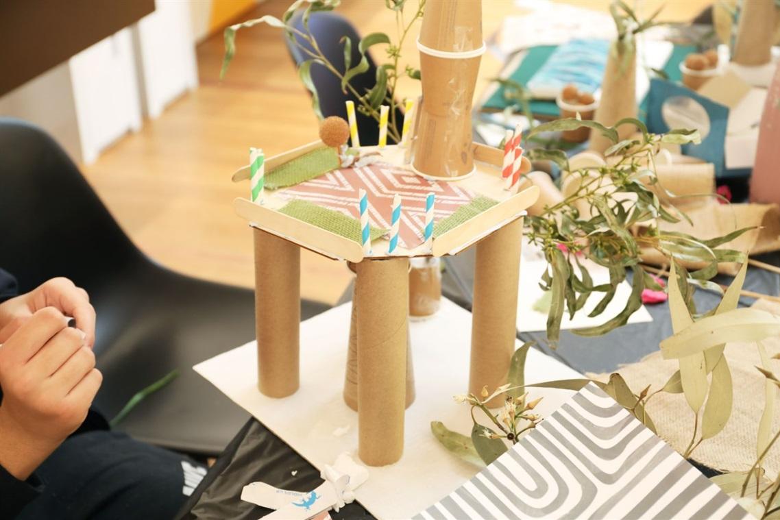 A child crafts a structure from cardboard tubes, paper, and leaves at a table. Bright colors and a zigzag pattern add playful creativity.