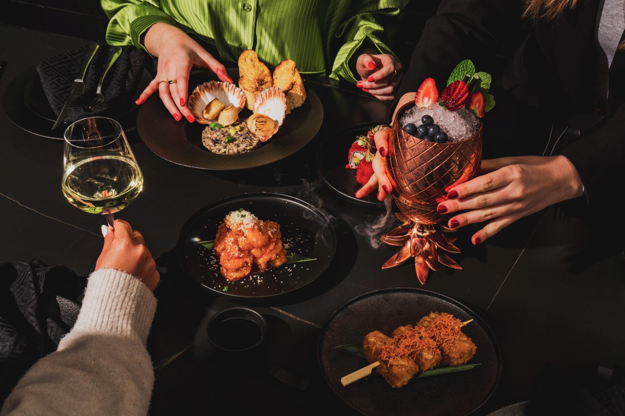 A stylish dinner setting with diverse dishes, including skewers and seafood. Three hands hold a cocktail, wine, and wearing a green blouse. Elegant and festive.