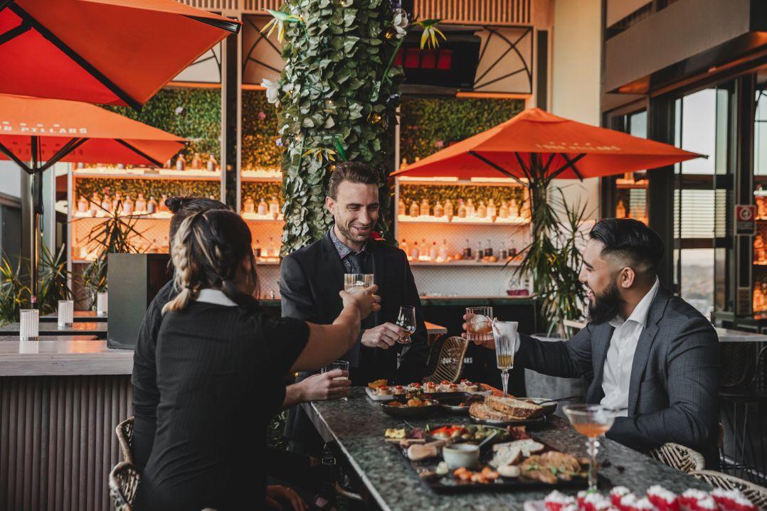 Three people in business attire enjoy drinks and appetizers at an upscale bar with greenery and orange umbrellas, sharing a cheerful toast.