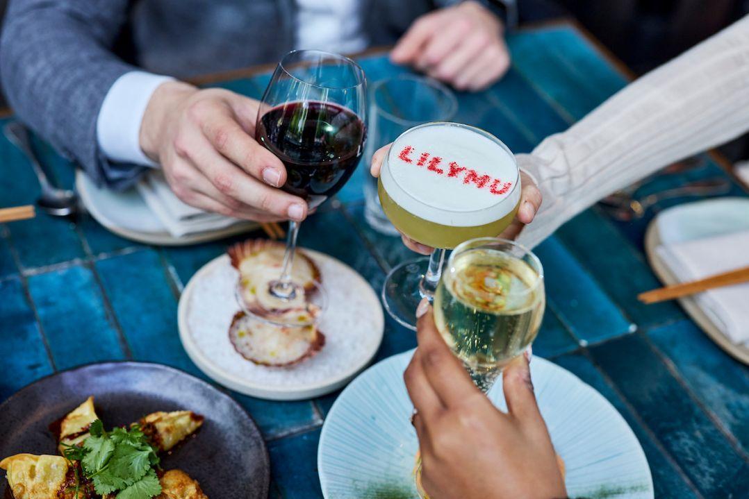 Three hands raise glasses in a toast over a blue table. Drinks include red wine, a cocktail with "LILYMU" on top, and champagne. Plates of food are visible.