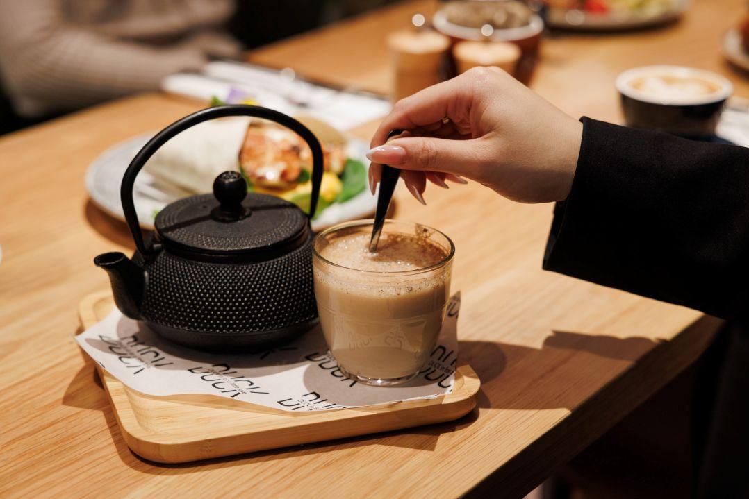 A hand stirs a frothy latte in a glass next to a black teapot on a wooden tray. Background includes a dining table with various food dishes.