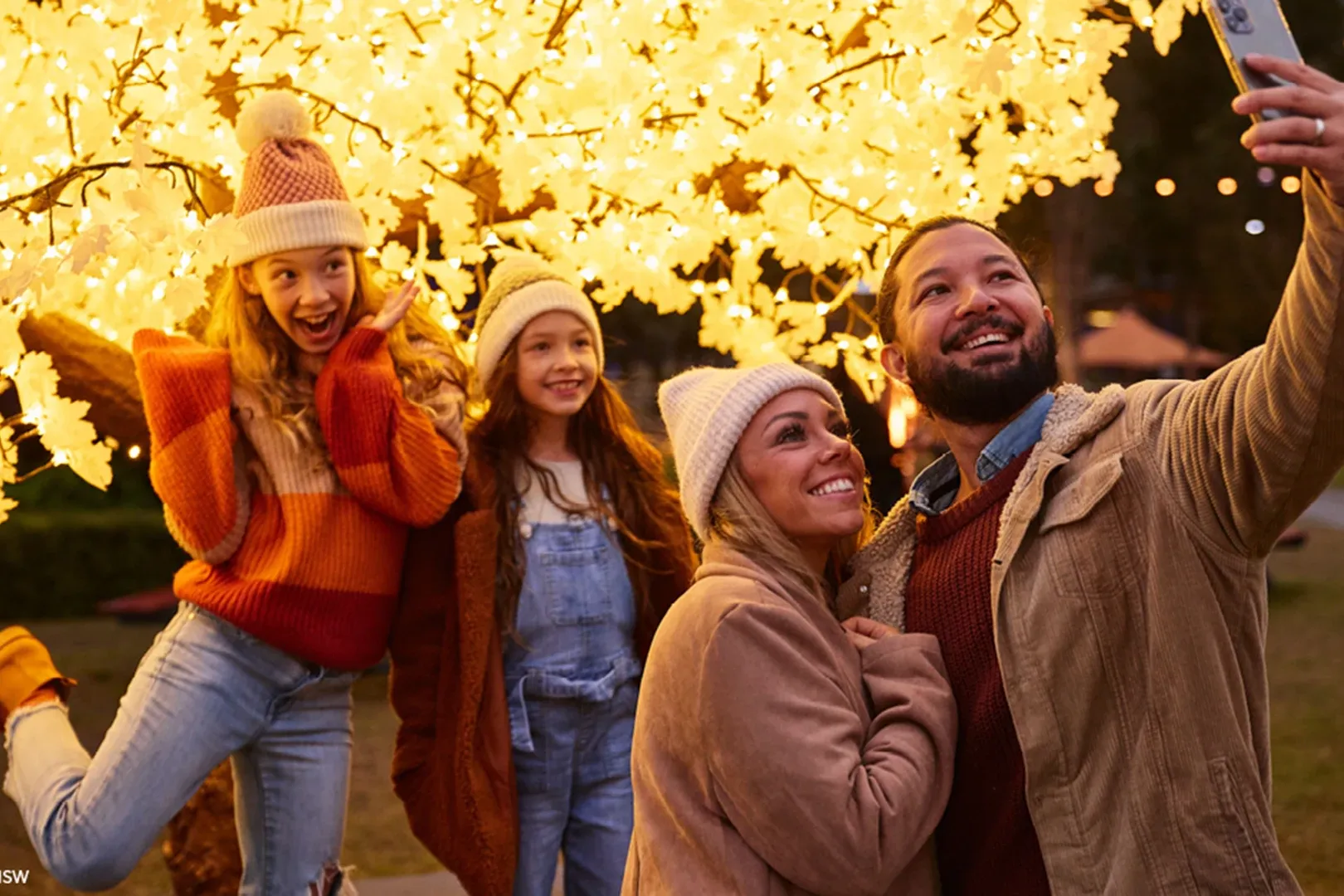 A joyful family takes a selfie under a glowing tree adorned with warm yellow lights at night. They wear cozy autumn clothes, expressing happiness and warmth.