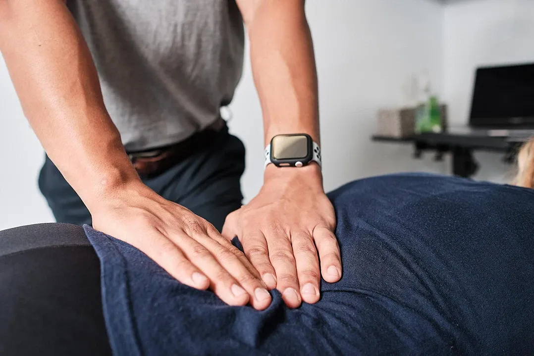 A person wearing a smartwatch performs a massage on someone lying face down, covered in a navy towel. The scene suggests relaxation and relief.