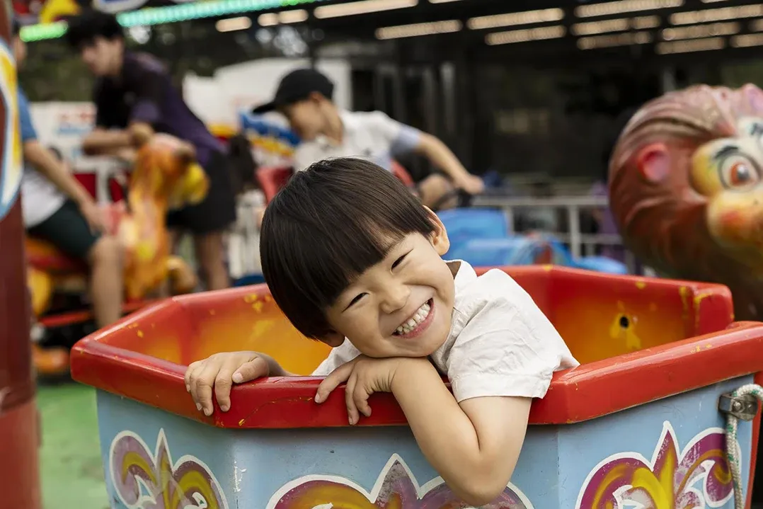 A joyful child with short black hair grins widely while leaning on the edge of a colourful carnival ride's seat. The background shows people enjoying the ride.