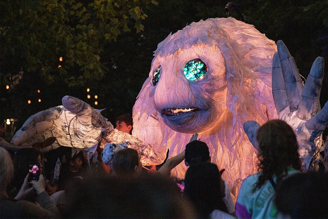 A large, glowing puppet resembling a friendly creature with bright, sparkling eyes is surrounded by a crowd. It's dusk, and the scene is festive and lively.