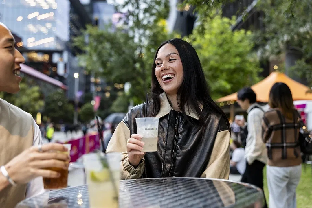 A woman holding a drink laughs with a friend at an outdoor event. They're seated at a round table, surrounded by greenery and modern buildings.