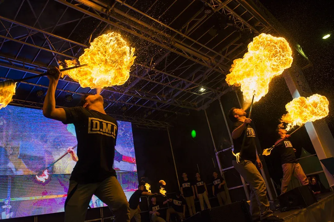 Fire performers on stage breathe flames under a metal framework, creating a dramatic and intense atmosphere. Audience stands captivated in the background.