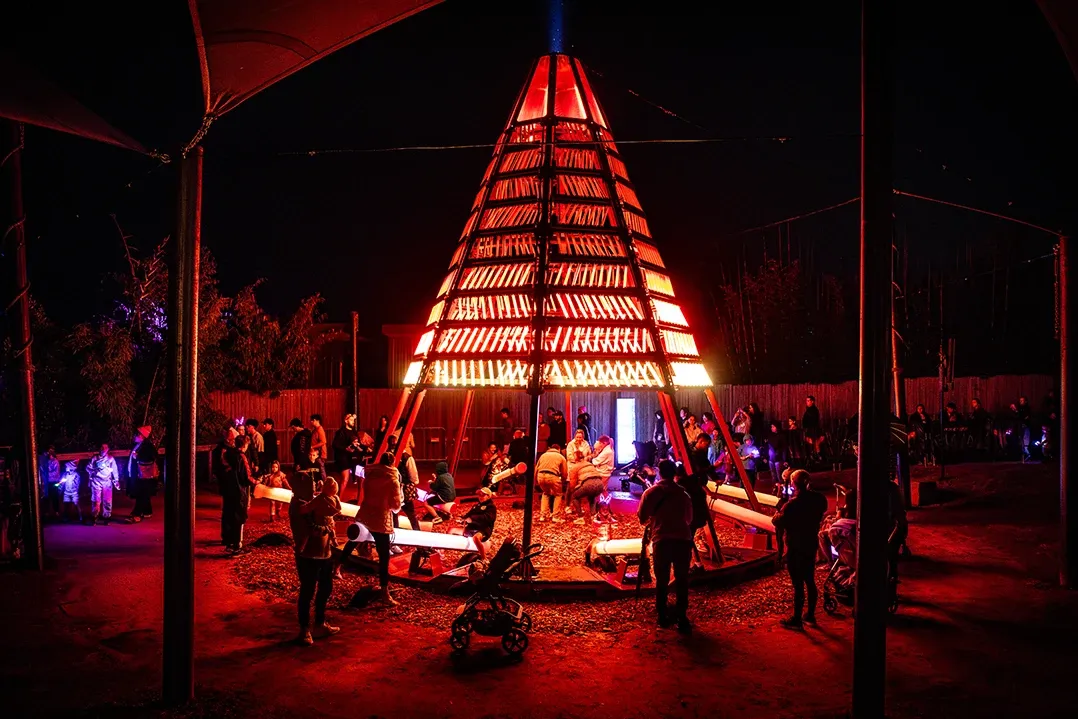 A large illuminated teepee surrounded by people at night. The structure glows red, creating a warm, festive atmosphere. Families and children gather around it.
