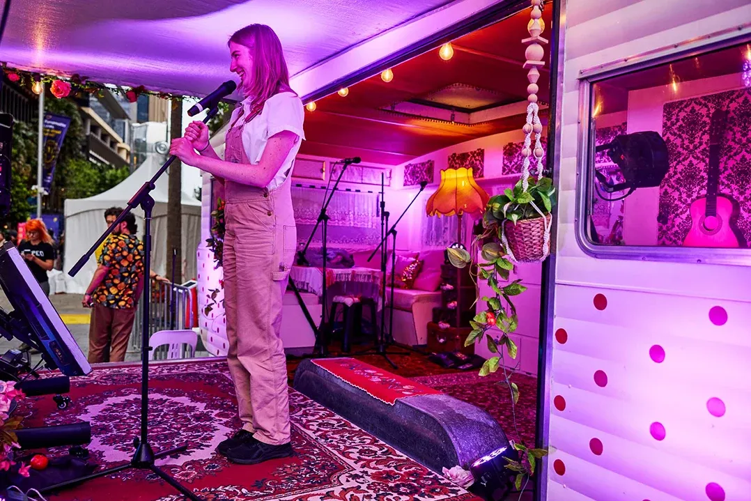 A woman in casual attire stands on a decorated stage inside a vibrantly lit caravan. She is holding a microphone and smiling, conveying a joyful atmosphere.