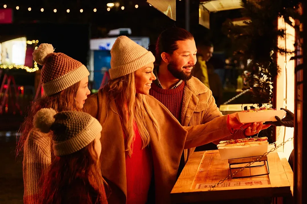 A joyful family of four, dressed in cozy winter clothing and hats, selects food from a brightly lit festive market stall. Warm atmosphere.