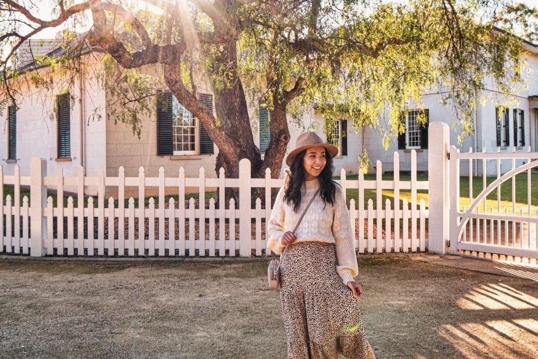A woman in a cozy sweater and leopard print skirt stands smiling by a white picket fence. Sunlight filters through trees, creating a warm, serene mood.