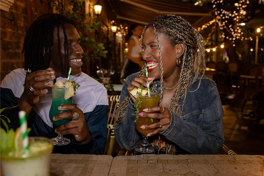 A smiling couple enjoys colorful cocktails at an outdoor cafe, decorated with string lights, creating a warm and festive evening ambiance.