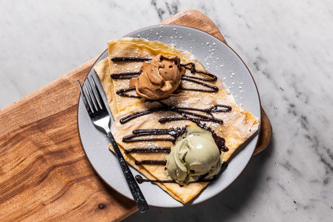 Crepe dessert with a scoop of brown and a scoop of green ice cream on top, drizzled with chocolate sauce, powdered sugar, fork on the side, on a wooden board.
