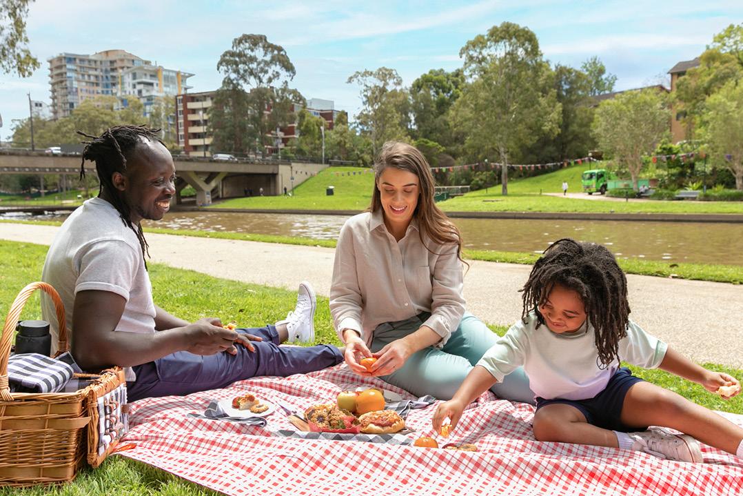 A family of three enjoys a picnic on a red-checked blanket by a riverside park. They're smiling, surrounded by food, under a clear blue sky.