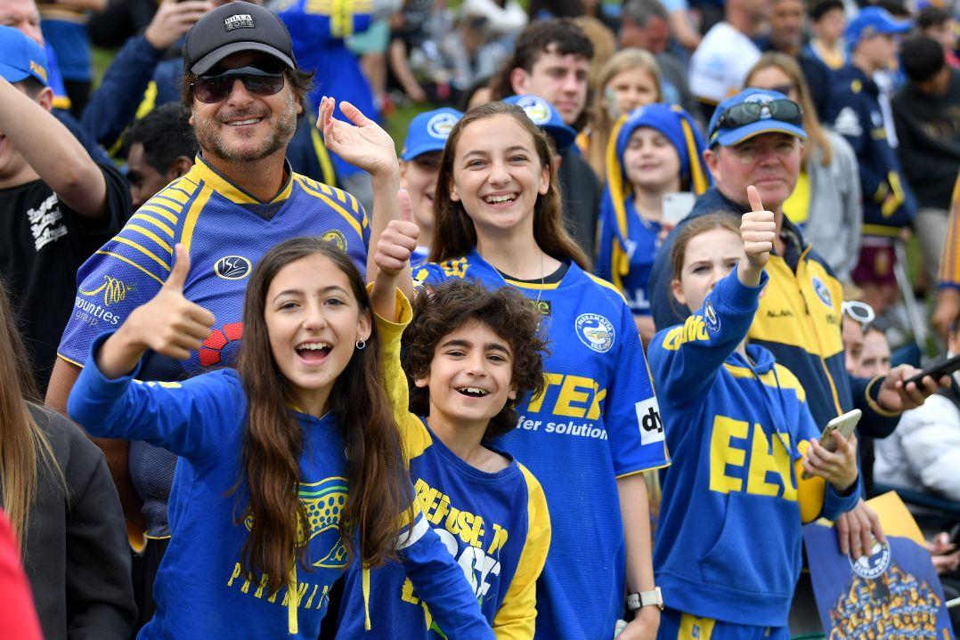 A group of enthusiastic fans wearing blue and yellow jerseys, some giving thumbs up and smiling, in a lively crowd at a sports event.