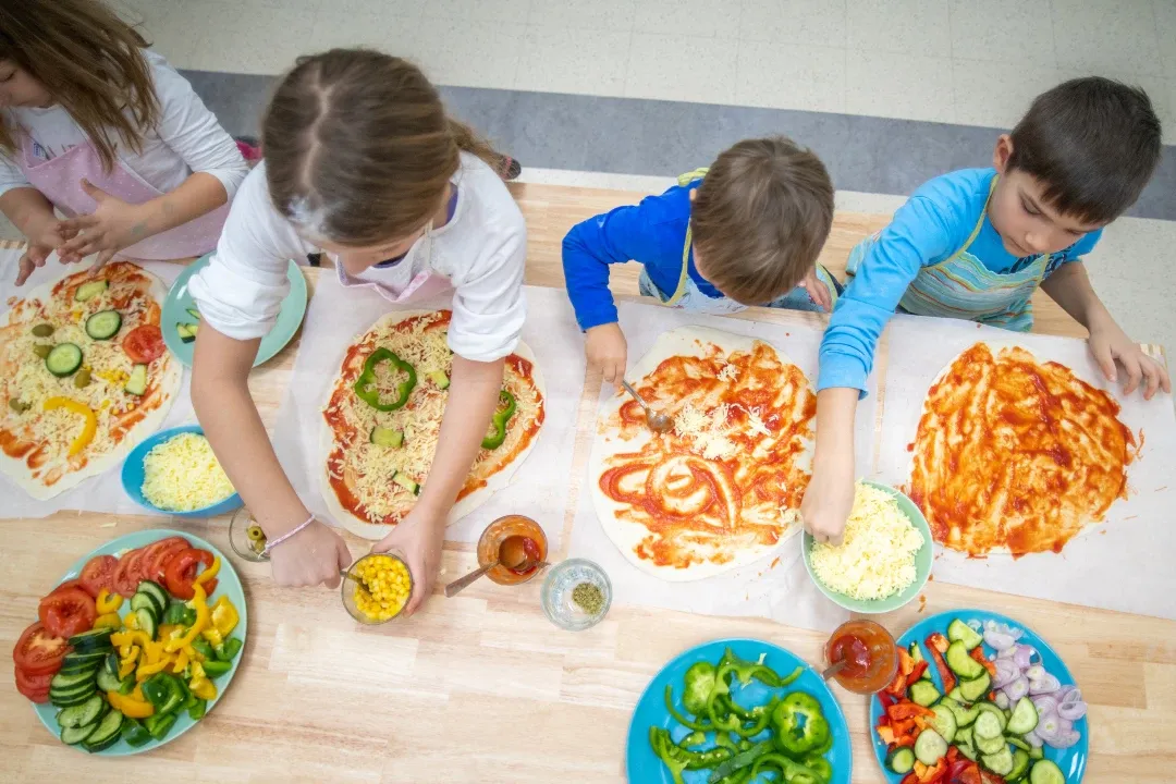 Children enthusiastically make pizzas at a wooden table. Toppings like cheese, bell peppers, and cucumbers are spread around. The scene is lively and creative.