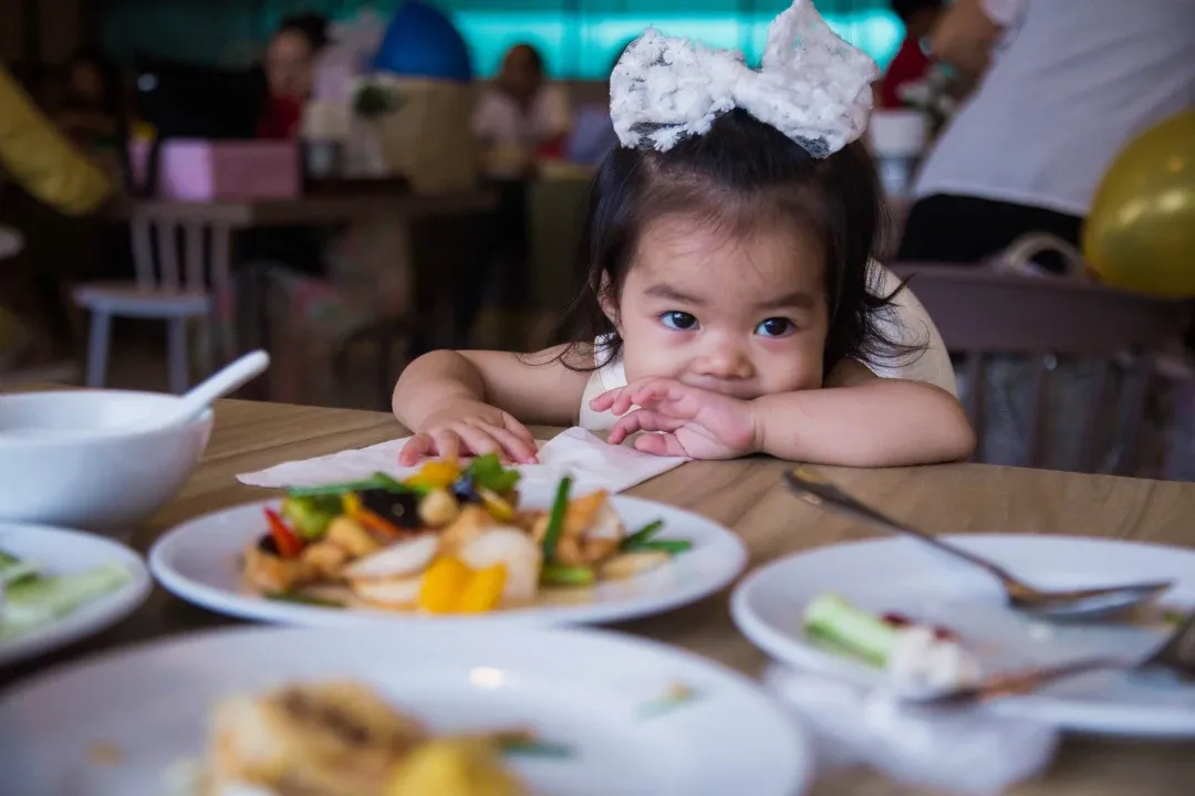 A toddler with a large white bow leans on a table filled with plates of colorful food, gazing curiously. The setting is a lively, warmly lit restaurant.