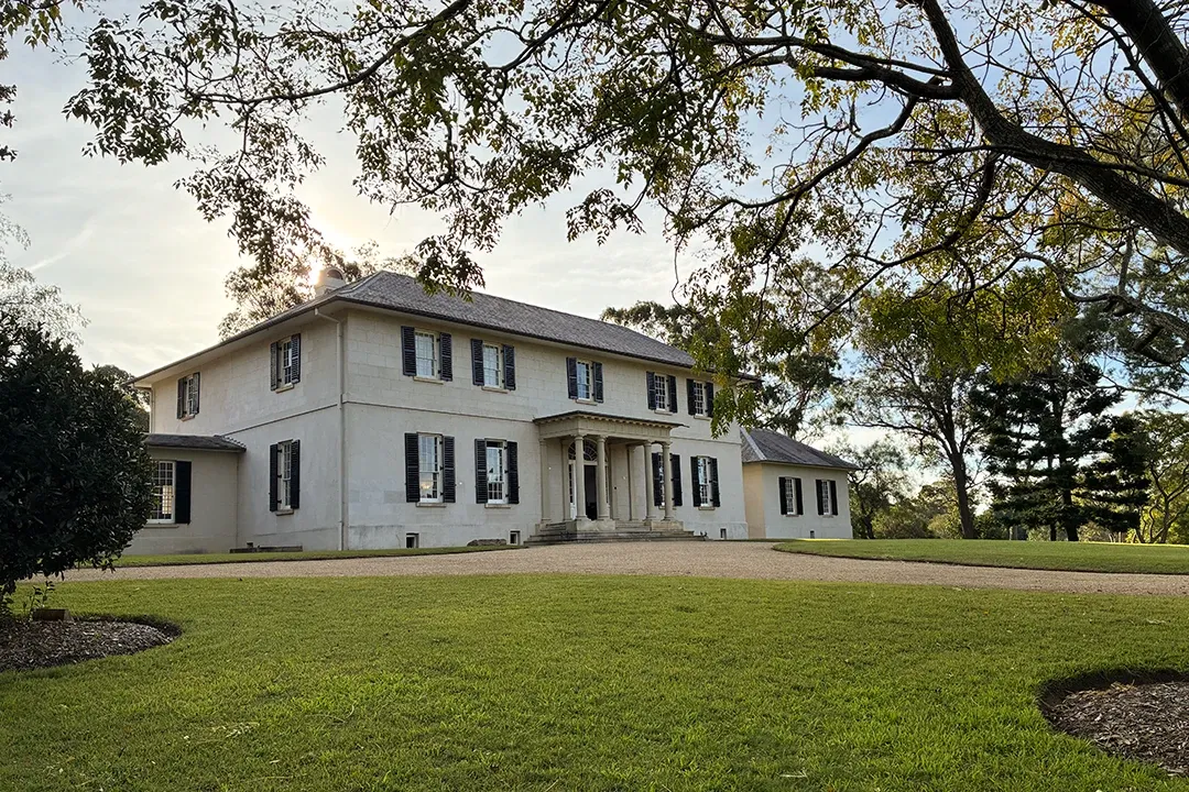 Large historic house with white walls and dark shutters, surrounded by lush greenery and expansive lawn under a clear sky; serene ambiance.