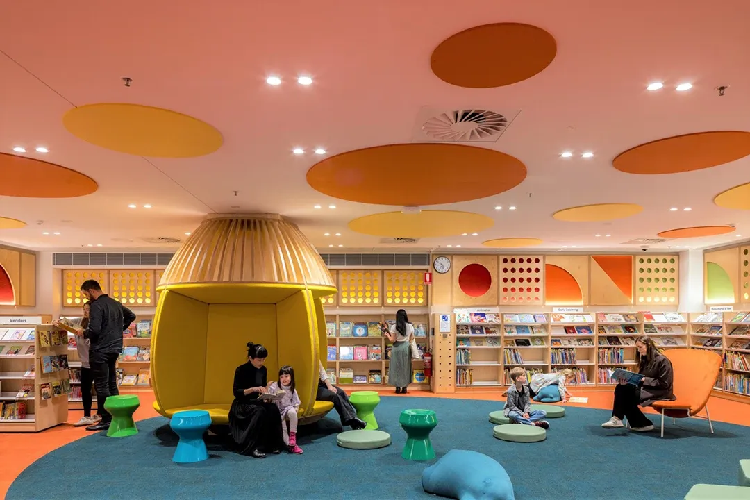 A cozy children's library with bright orange and yellow decor. Kids and adults sit on colorful seats reading, under whimsical ceiling designs.