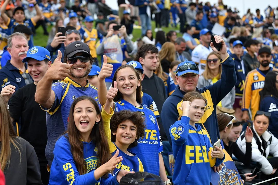 A joyful crowd wearing blue and yellow sports jerseys enthusiastically cheers and gives thumbs up, creating a lively and energetic atmosphere in a stadium.