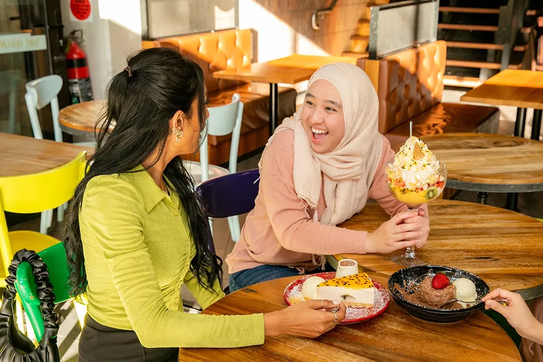 Two women are sitting at a wooden table in a cozy café, sharing dessert. They are smiling and appear to be enjoying their conversation.