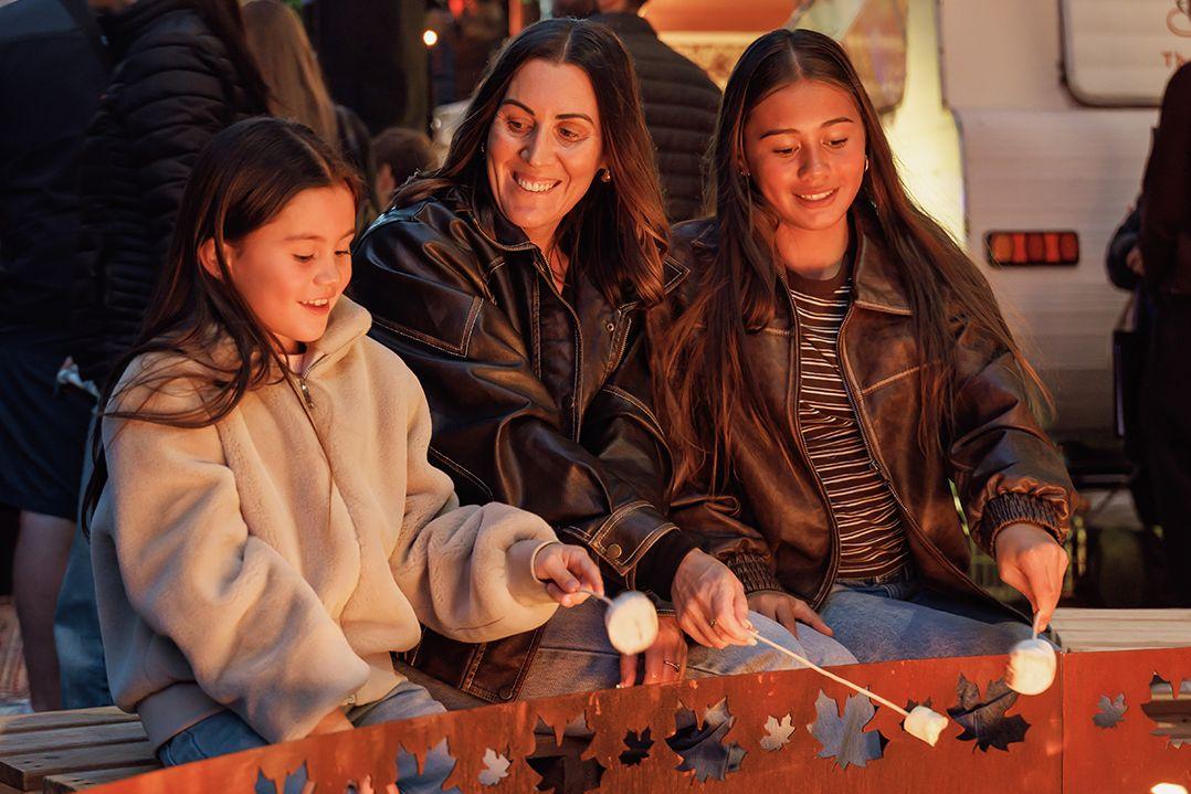 a woman and two children sitting by a bon fire with marshmallows on sticks