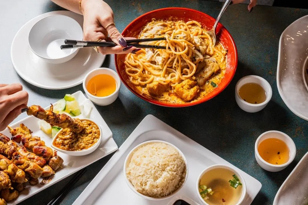 A vibrant Asian meal featuring a red bowl of noodle soup, skewered meat with peanut sauce, rice, and various dipping sauces on a dark table.