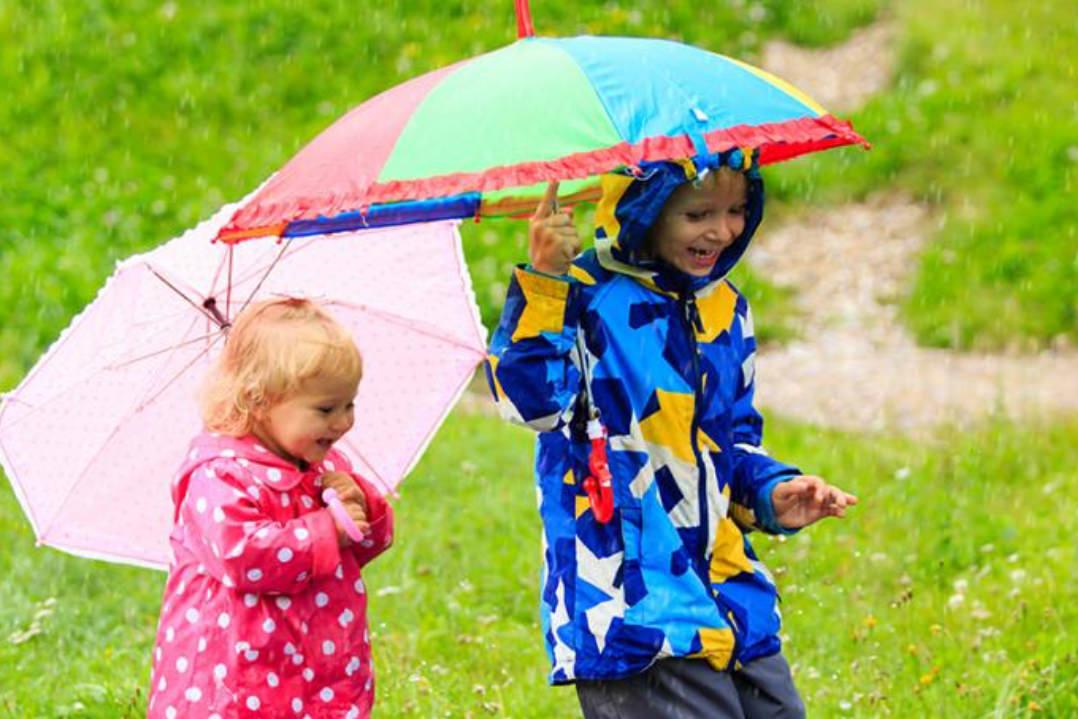 Two children in colorful raincoats laugh under vibrant umbrellas in a rainy green field. The scene conveys joy and playful adventure.