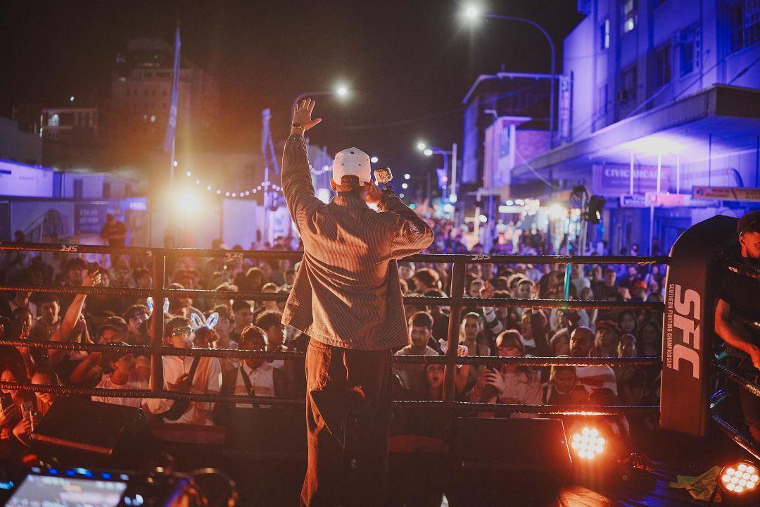 Performer raises both hands toward a cheering nighttime crowd from a lit stage, with city buildings, streetlights, and vivid purple-orange lights behind.
