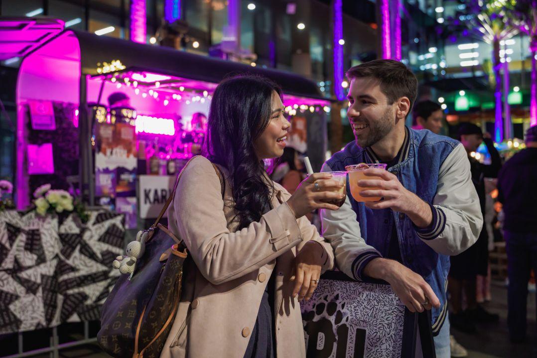 A smiling woman in a beige coat and a man in a blue varsity jacket toast with drinks in front of a brightly lit, purple-hued outdoor food stall.