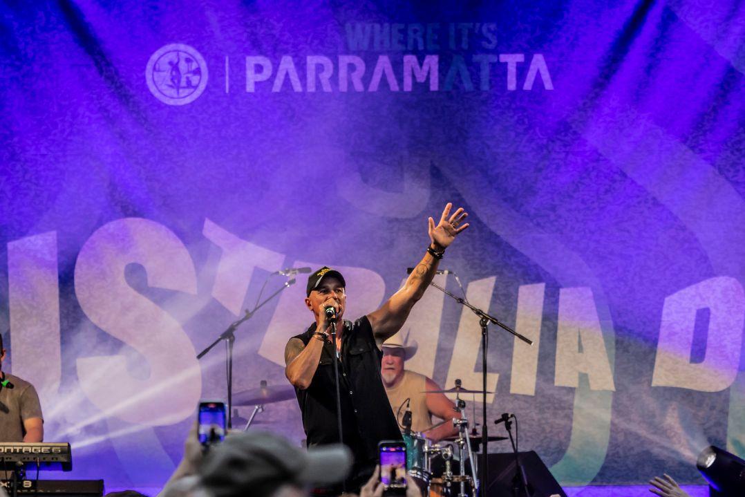 Singer performs into a microphone, raising one hand on a purple-lit stage with a drummer behind him and “Parramatta” signage overhead.