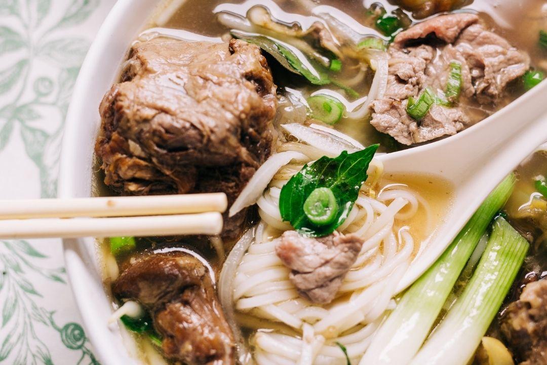 A close-up of a bowl of beef pho with rice noodles, tender meat chunks, green onions, and a fresh basil leaf, served with a white spoon and chopsticks.