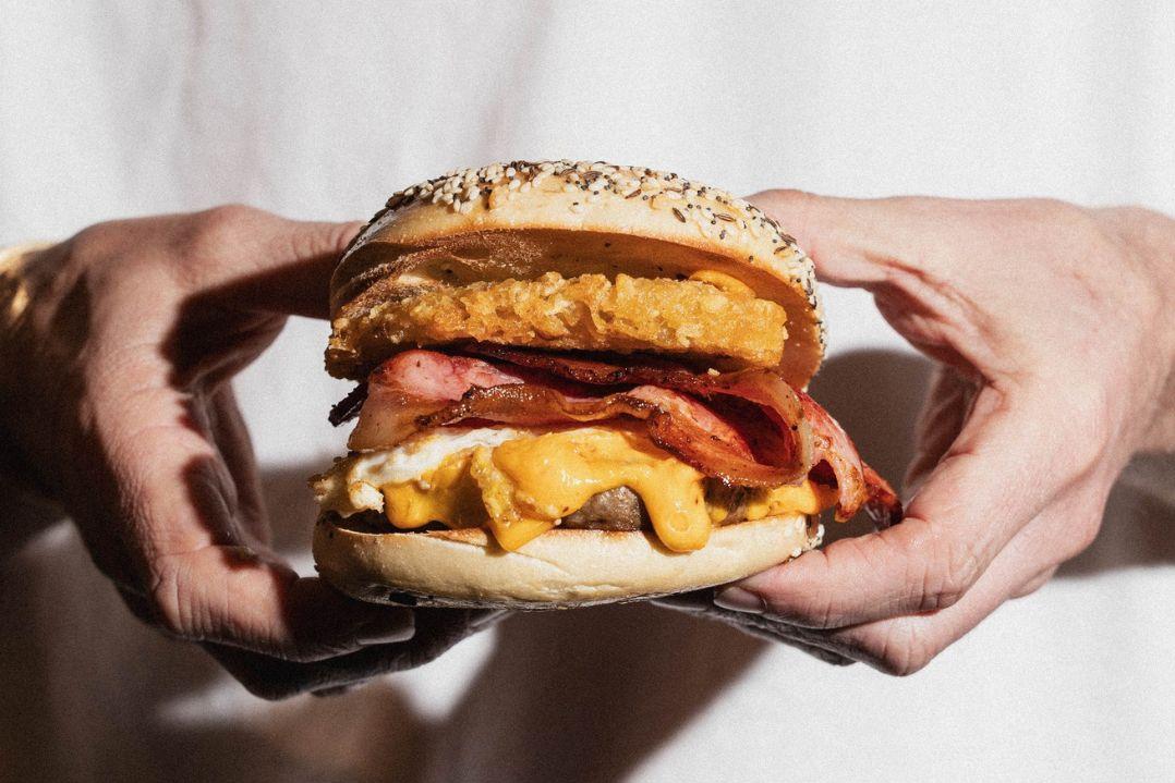 The toasted seeded bagel is filled with a fried egg, melted cheese, crispy bacon and a golden hash brown, with fillings layered generously between the bun against a plain light-coloured background.