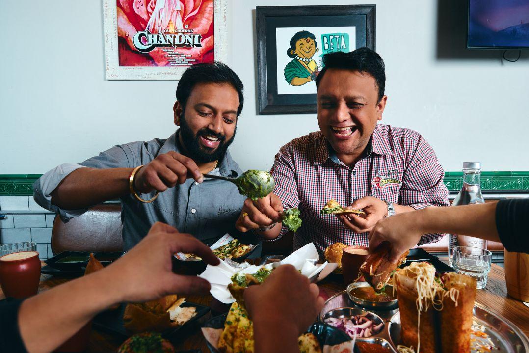 Friends laugh and share a colorful meal at a dining table, serving food from communal dishes in a warm, lively, joyful gathering.