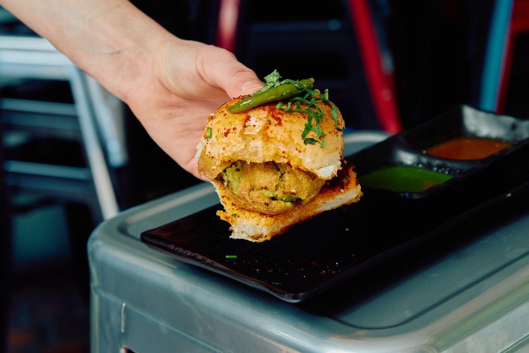 Close-up of a vada pav served on a black tray, featuring a spiced potato fritter inside a soft bread roll topped with herbs and green chilli.