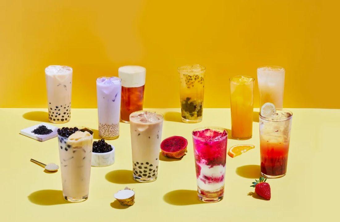 A variety of colorful bubble teas and fruit drinks in clear glasses arranged on a yellow surface with scattered fruit pieces and boba pearls.