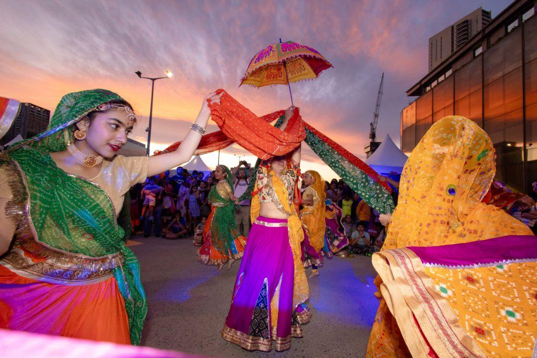 Women in vibrant traditional Indian attire dance joyfully outdoors in a lively festival setting, holding colorful scarves against a dramatic sunset sky.
