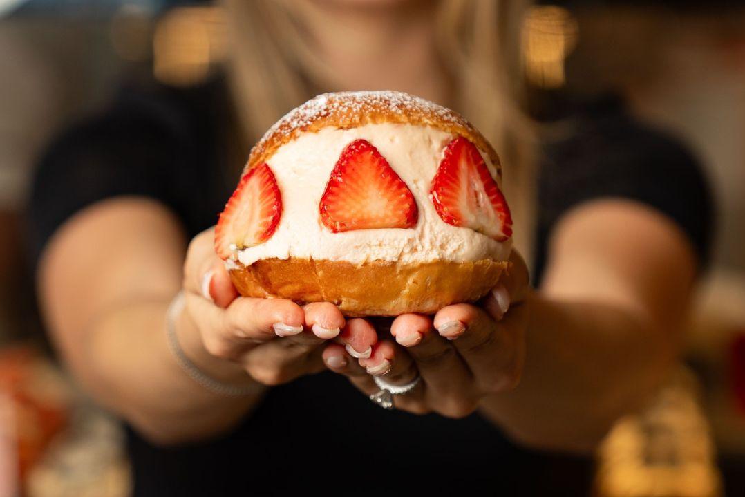 Person holding a large cream-filled doughnut topped with fresh strawberry slices and a dusting of icing sugar.