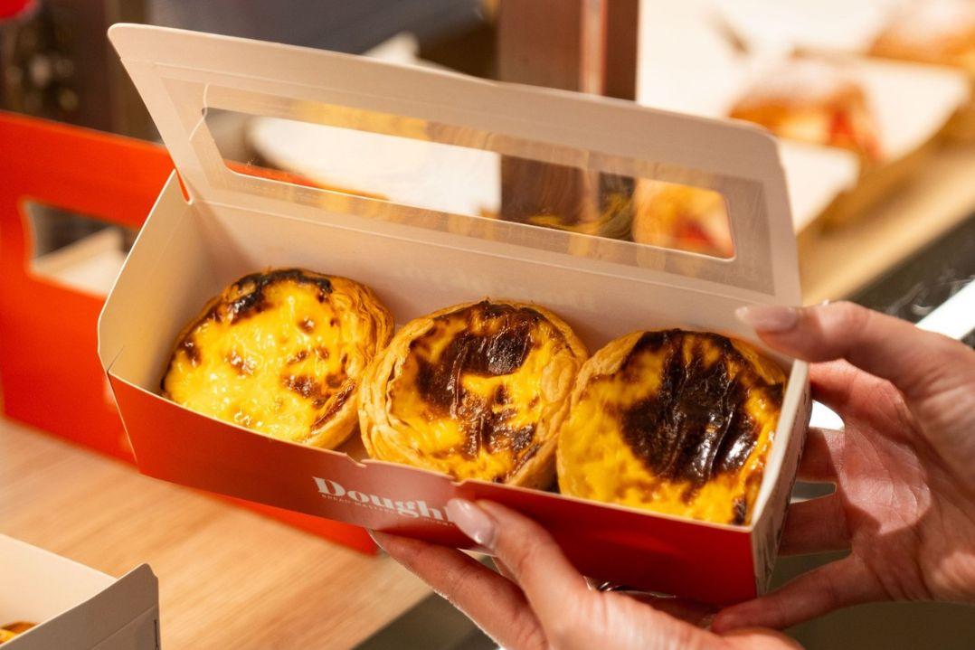Box of three Portuguese custard tarts with caramelised tops, held in two hands above a café counter.
